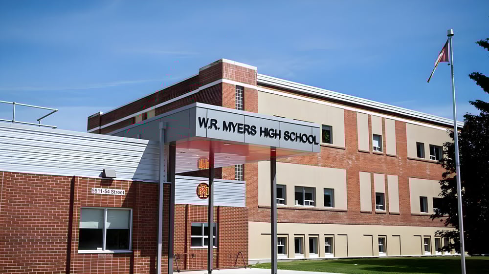 Das Gebäude der W.R Myers High School mit gut sichtbarem Schriftzug vor blauem Himmel und einem Fahnenmast im Vordergrund.