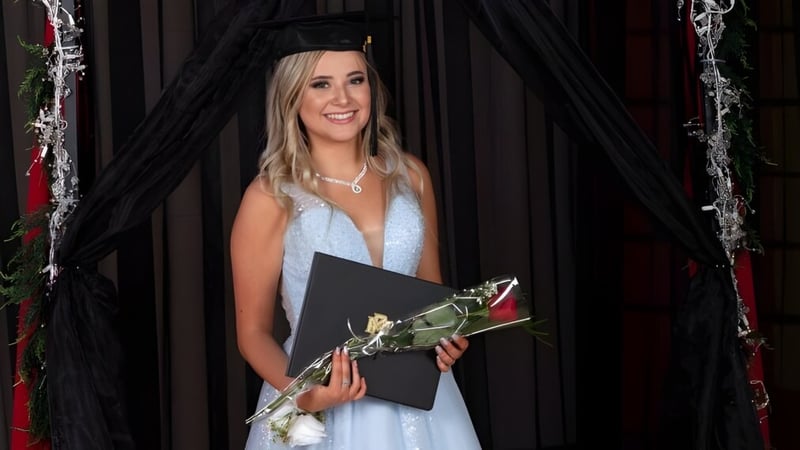 Eine Absolventin der W.R Myers High School steht mit blauem Kleid, Abschlussmütze, einem Diplom und einer roten Rose vor einem schwarzen Vorhang.