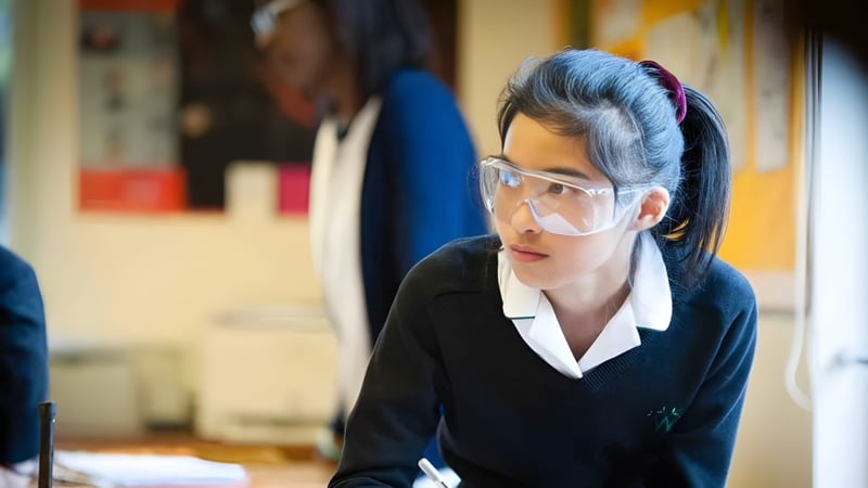 Eine junge Frau in schwarzem Anzug und Brille steht konzentriert in einem unordentlichen Raum der Wychwood School.