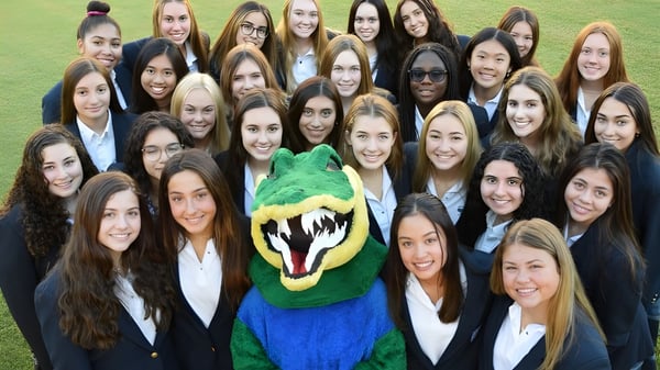 Eine Gruppe junger Frauen in formeller Kleidung steht auf einem Rasen mit dem grünen und blauen Maskottchen der Xavier College Preparatory High School.