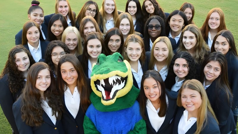 Eine Gruppe junger Frauen in formeller Kleidung steht auf einem Rasen mit dem grünen und blauen Maskottchen der Xavier College Preparatory High School.