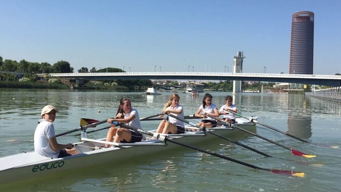Eine Gruppe von Schülerinnen und Schülern der Yago School rudert auf einem ruhigen Fluss mit Brücke und hohem Gebäude im Hintergrund.