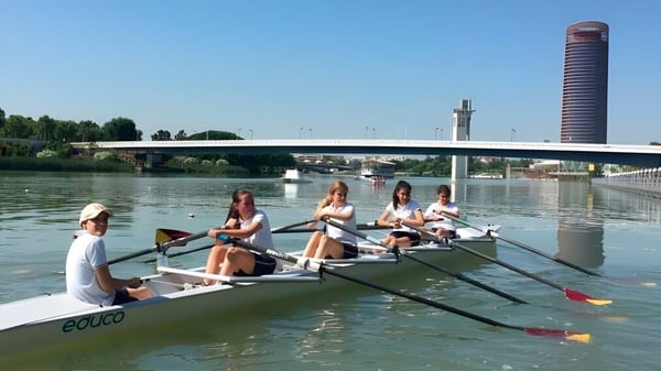 Schüler der Yago School rudern auf einem Fluss mit Brücke und hohem Turm im Hintergrund.