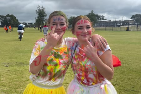 Zwei Schülerinnen des Yanchep Secondary College tragen bunte Tie-Dye-Shirts und machen im Freien Grimassen mit Handgesten.