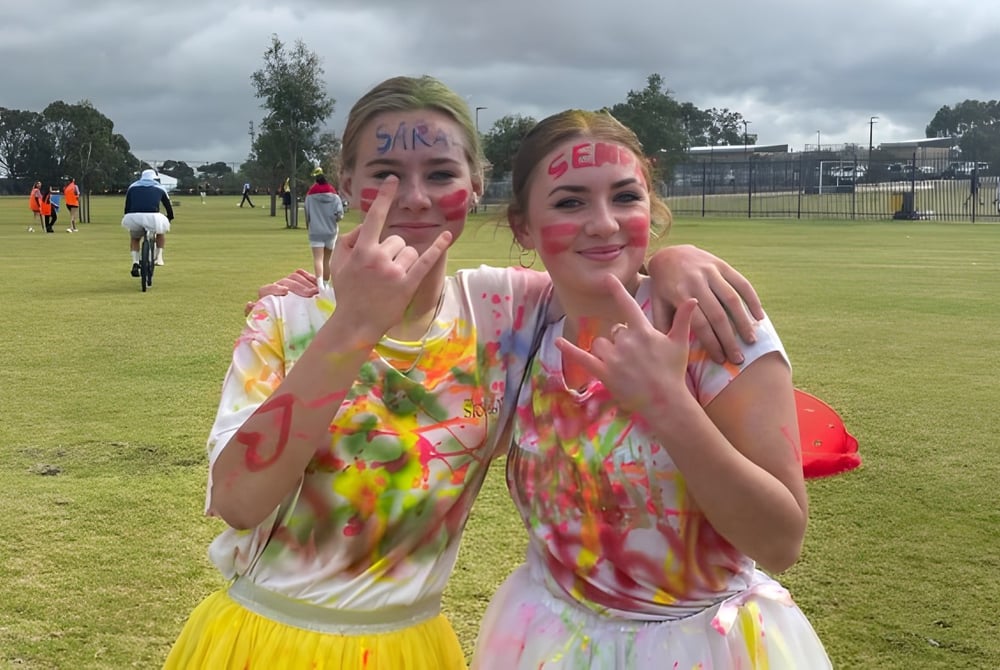 Zwei Schülerinnen des Yanchep Secondary College tragen bunte Tie-Dye-Shirts und machen im Freien Grimassen mit Handgesten.