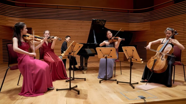 Eine Gruppe von Musikschülern der Yehudi Menuhin School tritt in formeller Kleidung auf einer Bühne mit einem Flügel und Holzvertäfelung auf.