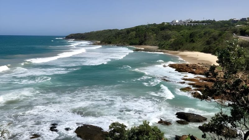 Eine Küstenlandschaft mit Sandstrand, Wellen und bewachsener Klippe nahe der Yeppoon State High School.