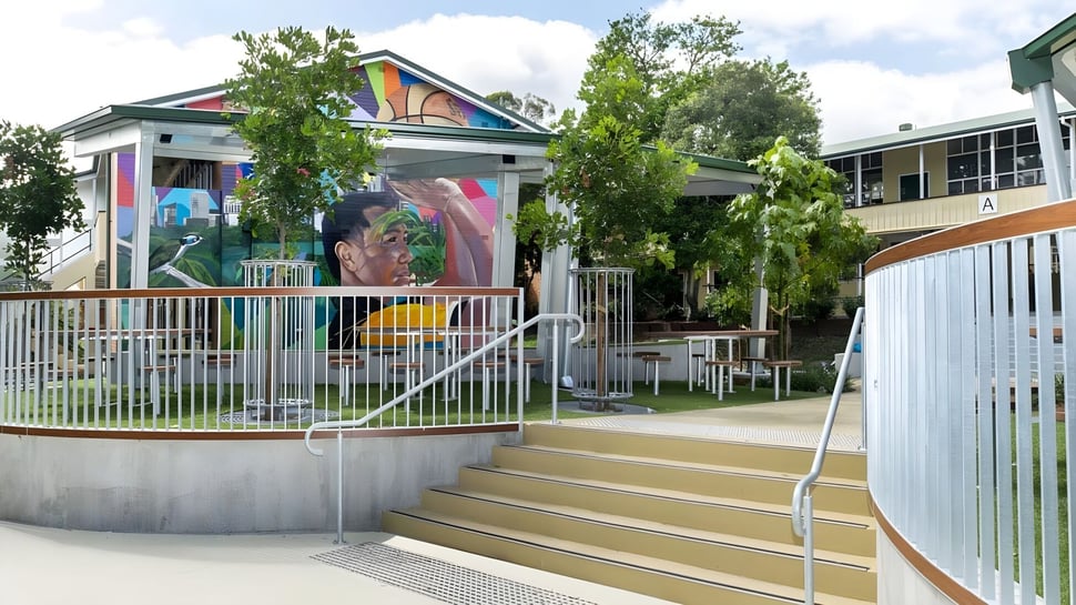 Das bunte Wandbild an der Fassade der Yeronga State High School ist von einem umzäunten Bereich mit Treppen und grüner Umgebung umgeben.