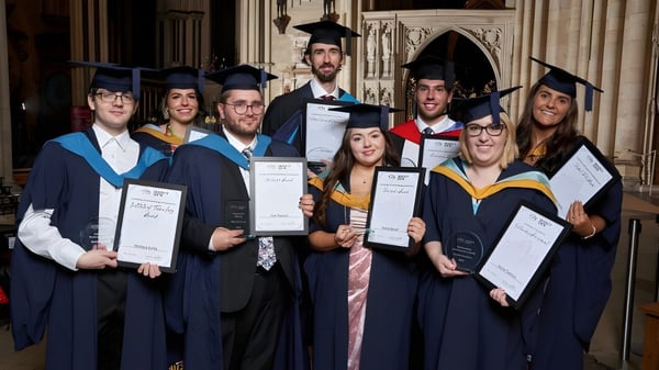 Eine Gruppe von Absolventen des York College steht gemeinsam in akademischer Kleidung in einem prächtigen Innenraum und hält ihre Diplome.