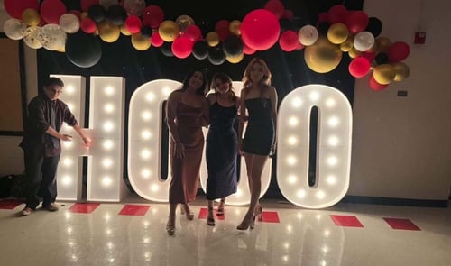 Drei Mädchen mit Kleidern in der Mitte und ein Junge links posieren vor LED Buchstaben und Ballons über ihnen