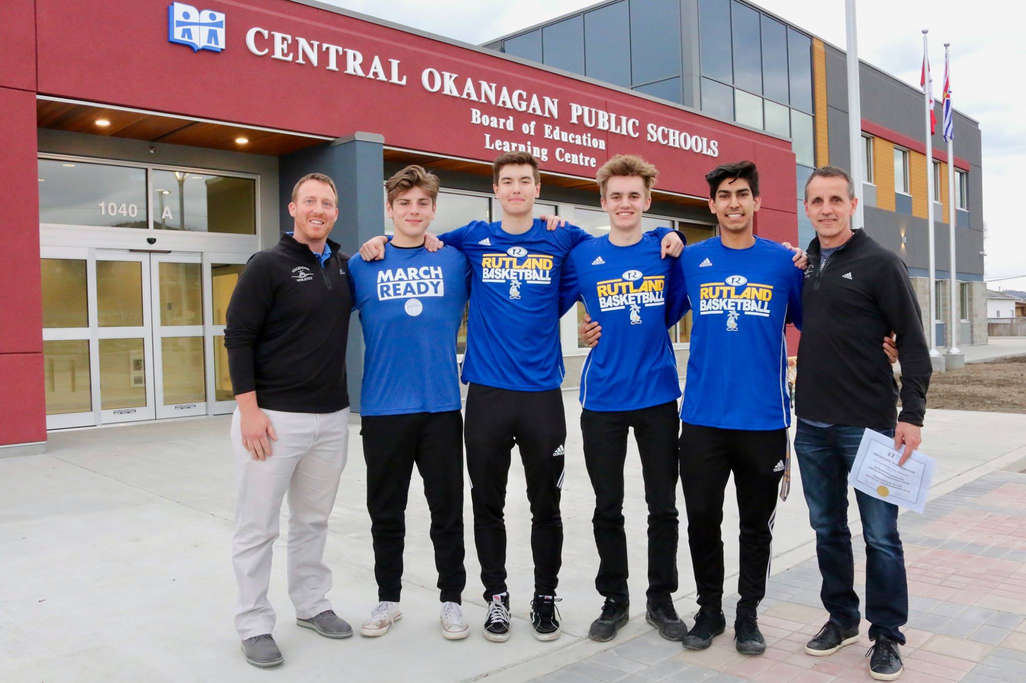 Eine Gruppe von Schülern steht mit zwei Lehrern in Basketball-Mannschaftstrikots vor dem Schulgebäude einer High School in Kanada