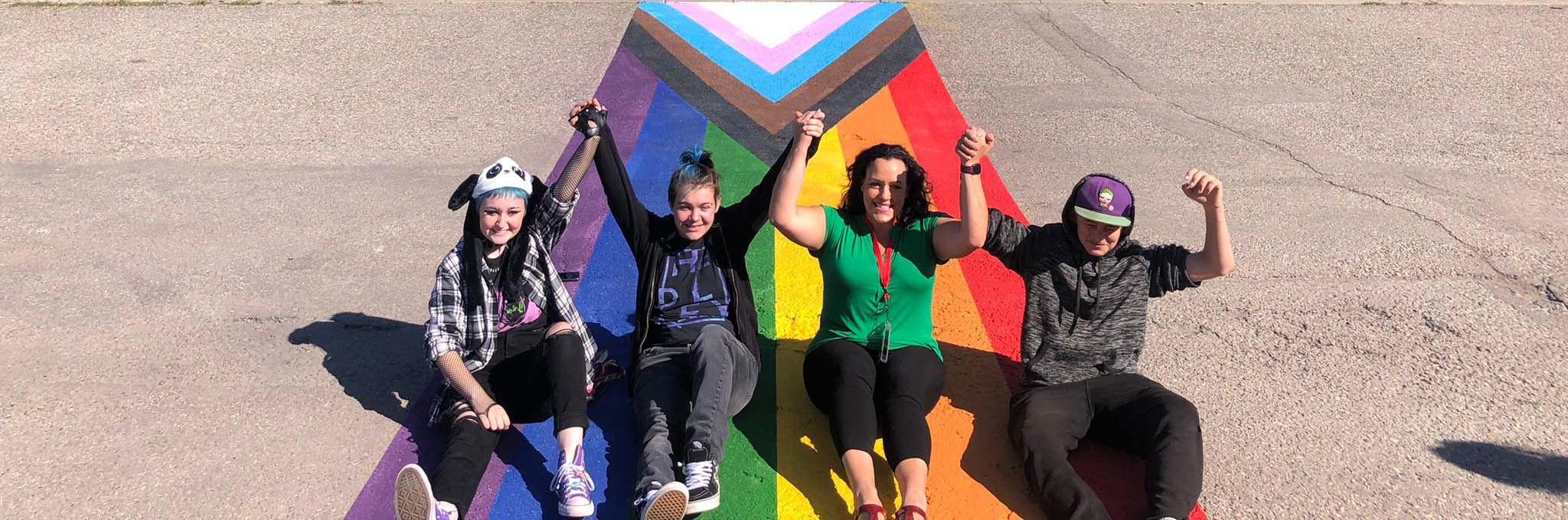 Eine Gruppe von Schüler:innen sitzen mit einer Lehrerin auf einem mit einer Pride-Flagge bemalten Asphalt-Weg