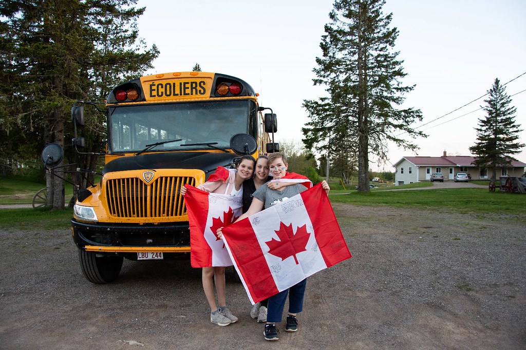 Drei Teilnehmer:innen stehen vor einem Schulbus und halten zwei Kanada Flaggen